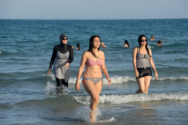 Nők a tengerparton. A balra lévő burkiniben, a teljes testet fedő, muszlim nőknek tervezett fürdőruhában - fotó: Fethi Belaid / AFP